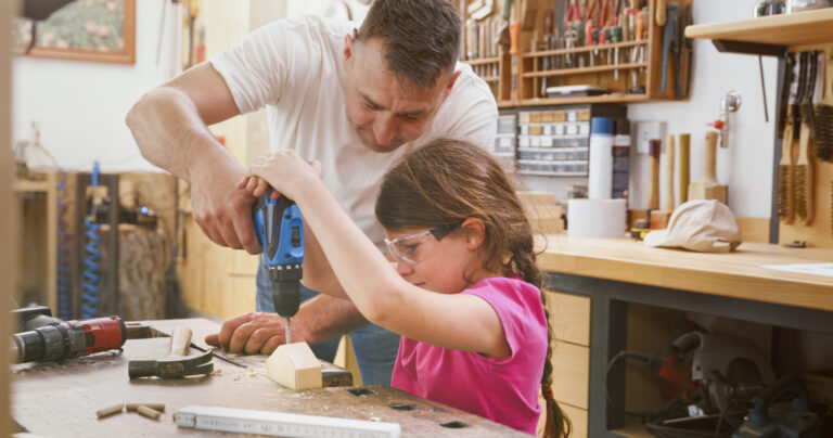 Father helping his daughter drilling hole on model wooden house in workshop.